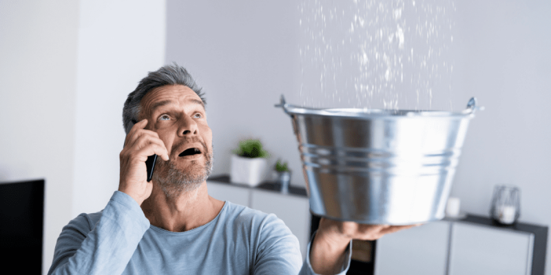 A man holding a bucket to catch water from a roof leak while talking on the phone to schedule an emergency roofing service.